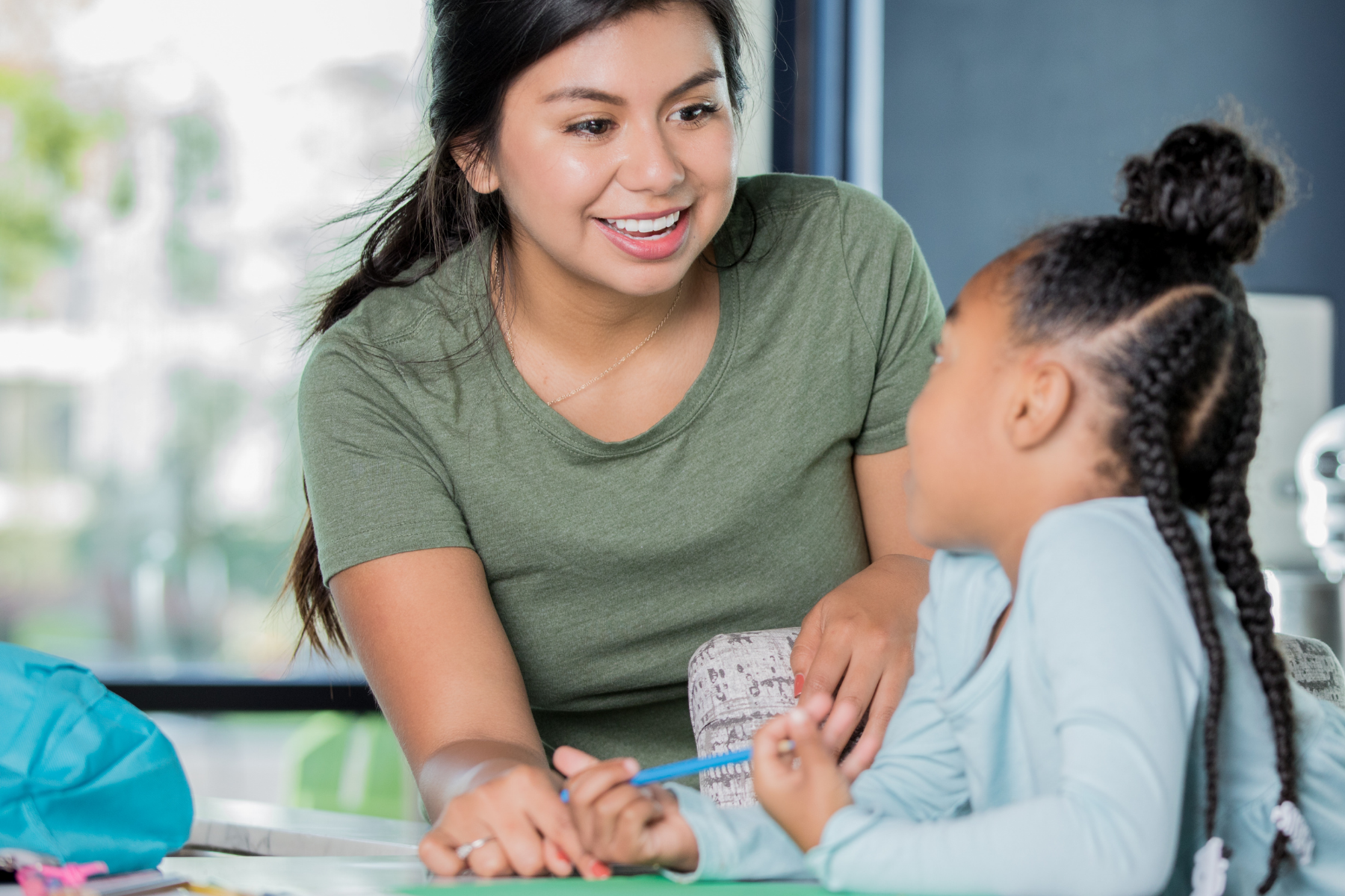 A smiling woman in a green t-shirt leans in to talk with a young girl sitting at a table. The girl has her hair in braided pigtails and a top bun and holds a blue pen over a green piece of paper. They are working at a white table with a large window in the background.